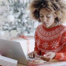 Young trendy African woman doing Xmas shopping online sitting in front of the decorated Christmas tree at home entering her card details on the computer to buy an mlm product